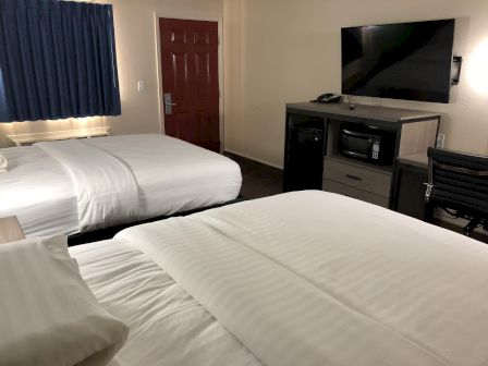 The image shows a hotel room with two beds, a TV, a microwave, a desk, a chair, and a window with blue curtains. The door is brown.