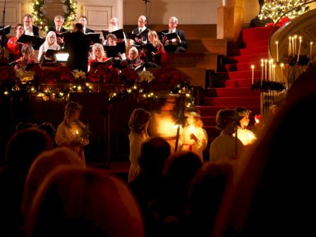 The image shows a choir performing in a decorated church, with women holding candles and an audience watching. Christmas decorations are visible.