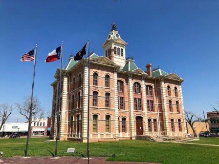 A historic courthouse with a clock tower and green roofs stands amid flags, under a clear blue sky, surrounded by a manicured lawn.