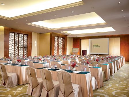 The image depicts a conference room set up for a meeting or seminar, with rows of chairs and tables, a projector screen, and elegant decor.