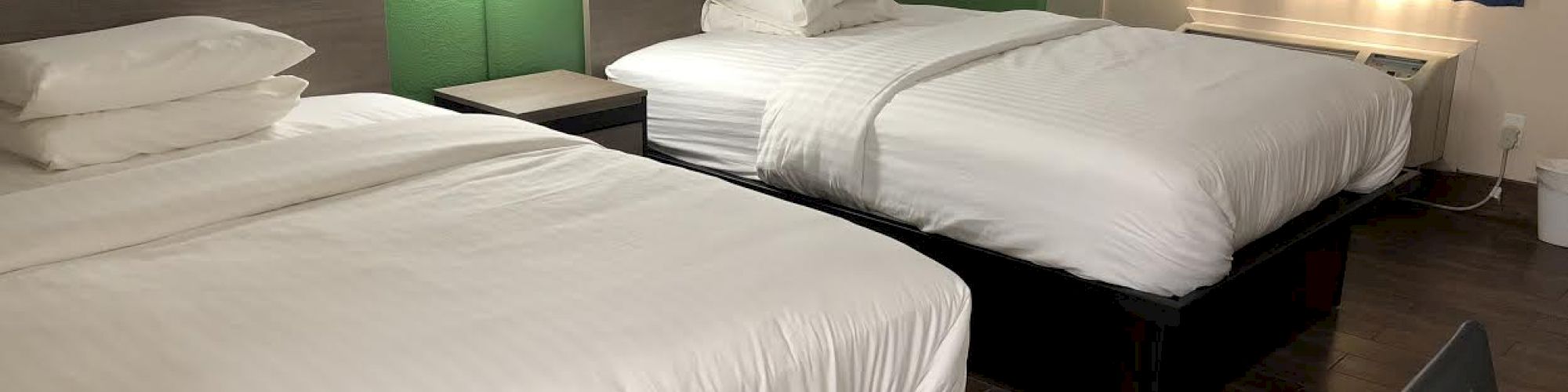 The image shows a hotel room with two neatly made double beds, a lamp on the wall, a window with blue curtains, and a wooden floor.