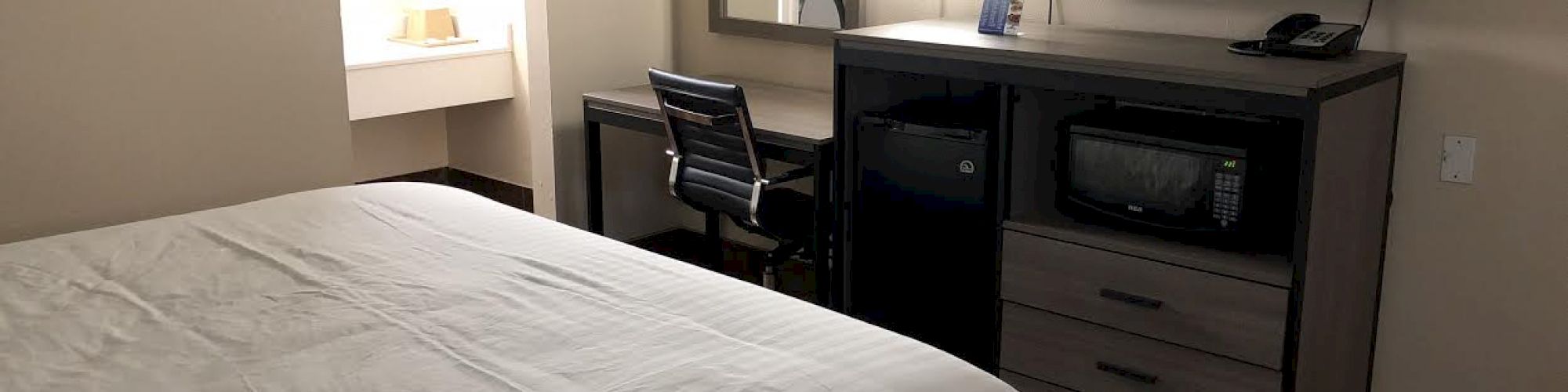 A hotel room with a bed, a desk, a chair, a wall-mounted TV, a dresser, a phone, a microwave, and a mirror.